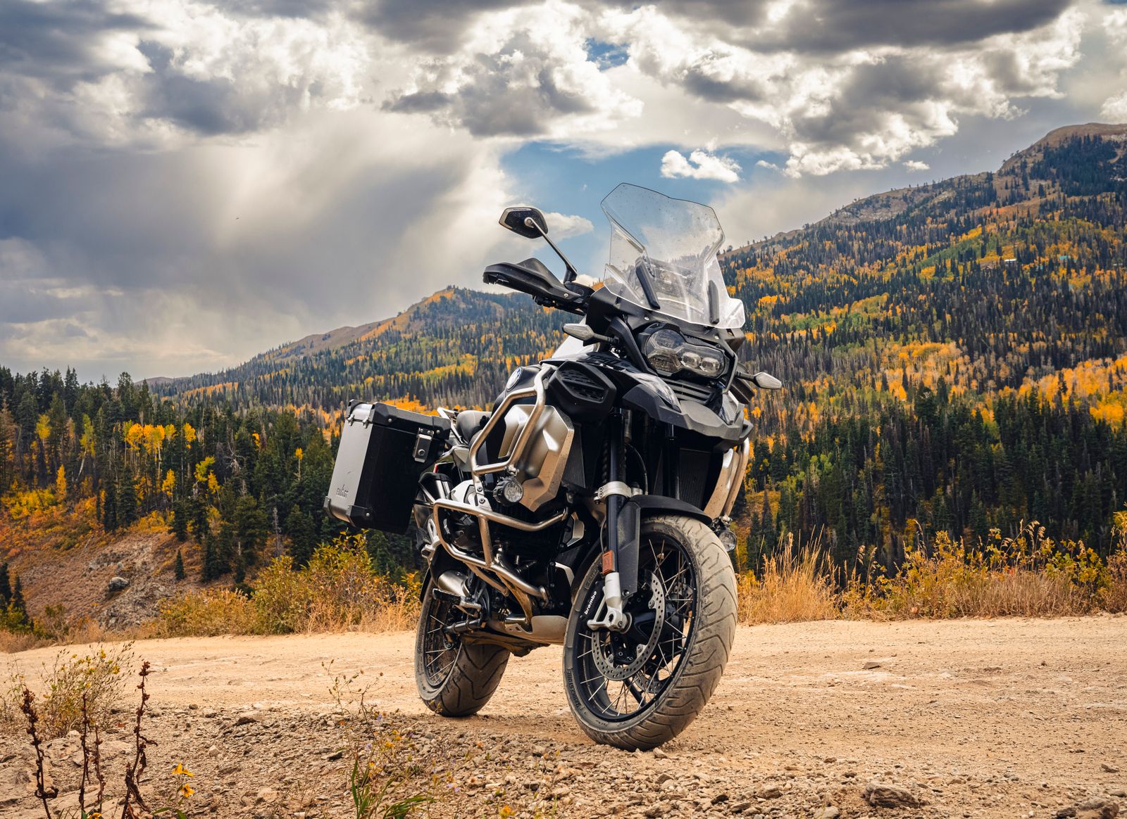 A parked adventure touring motorcycle with side panniers, overlooking a mountain forest with autumn colors under a cloudy sky