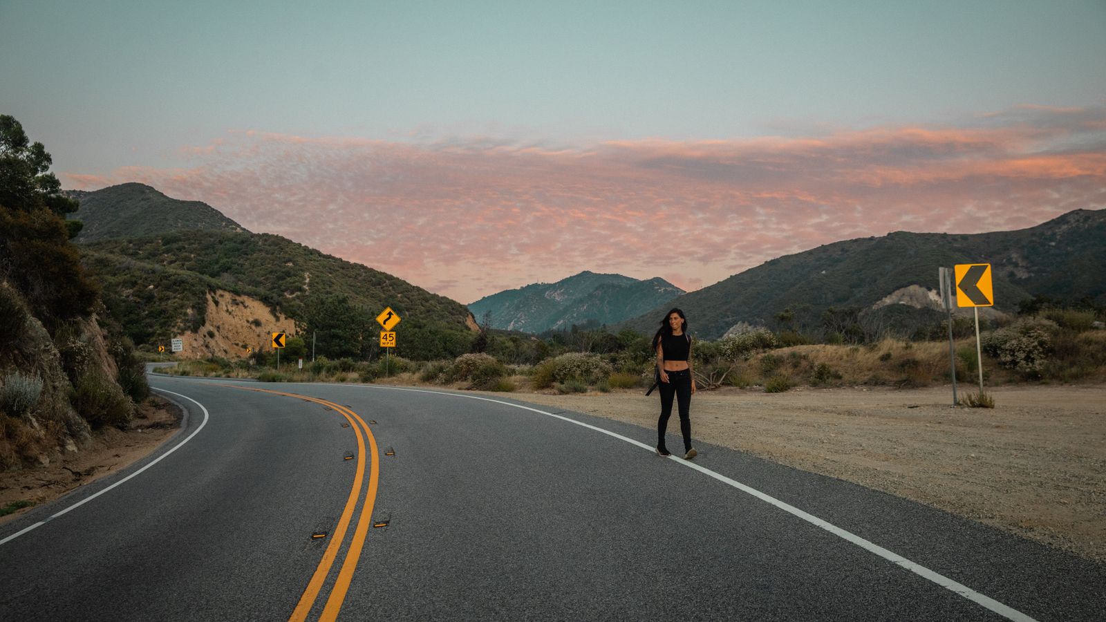 A lone traveler standing on an empty curving mountain road at dusk